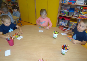 Przy stoliku siedzi troje dzieci. Dwoje z nich układa kredki na stole według wzoru na kartce. Jeden chłopiec spogląda w stronę pani.